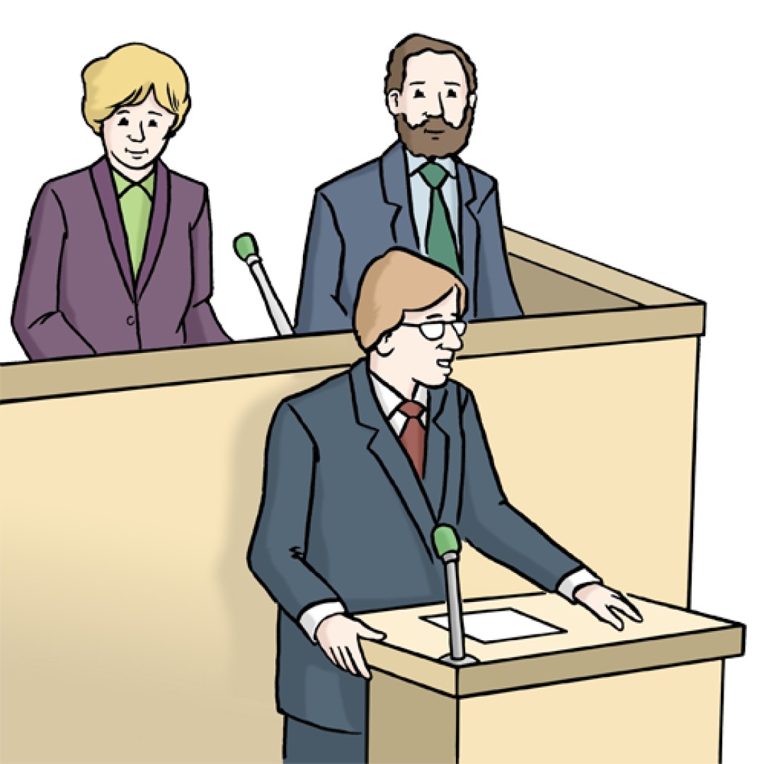 Das Bild zeigt 3 Menschen. Ein Mann hält eine Rede und spricht in ein Mikrofon. Eine Frau und ein Mann sitzen hinter dem Mann und hören zu. 